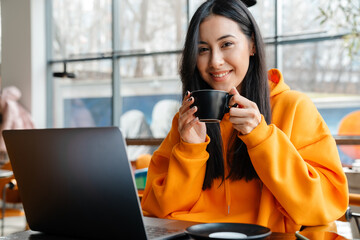 Smiling asian woman working with laptop while drinking coffee in cafe