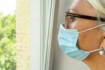 Fototapeta premium Sick caucasian female in glasses and medial mask gazing through to the window. Quarantined person indoors. Serene mornings. Avoiding social contact..