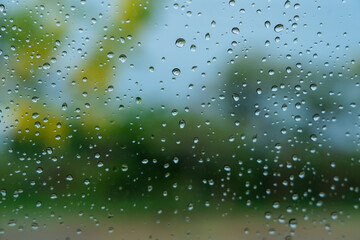 Rain droplets on surface of car glass with blurred green nature background and flowers through window glass of car. Freshness after rain. Soft focus, Selective focus on waterdrops. Natural background