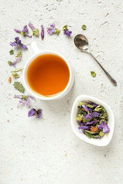 Herbal tea - thyme, green anise, ground ivy, pine bud, mallow flowers in a white cup and a tea spoon on a light gray table, top view with copy space