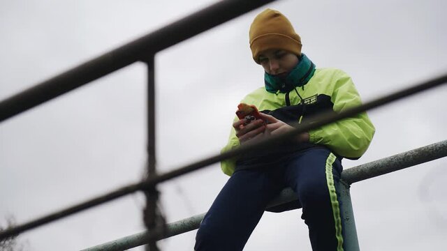 The boy sits on the metal bars on the playground and looks at the phone. Slow motion