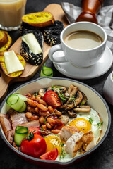English breakfast in pan with fried egg, beans, tomatoes, mushrooms, bacon and coffee on dark background. vertical image Top view