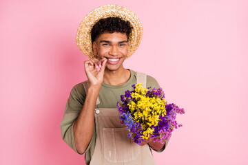 Photo of flirty florist guy hold bouquet wildflowers wear straw hat beige overall isolated pink color background