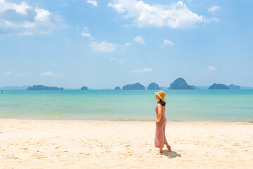 young woman wearing white dress standing on the beach and looking at blue sea in sunny day during vacation