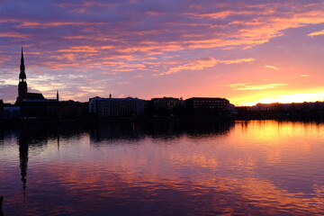 Fototapeta na wymiar Dawn over the Daugava in Riga, the sun illuminates the river, clouds are reflected in the water, the silhouette of Peter's Cathedral is visible