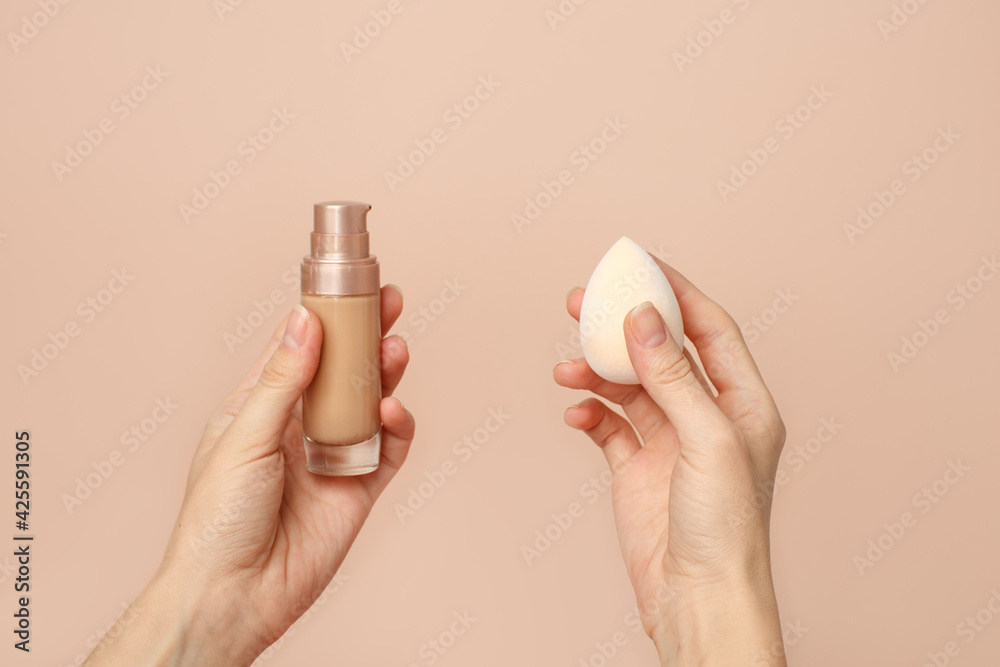 Wall mural closeup of female hands showing bottle of cosmetic foundation or concealer and sponge on neutral bei