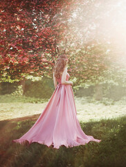 Spring woman walks in blooming sakura tree garden. Medieval long-haired girl, mysterious blonde lady in long pink satin silk elegant vintage dress. Bright fairy image, art photography, back rear view