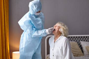 Doctor in protective suit taking throat and nasal swab from elderly female patient to test for...