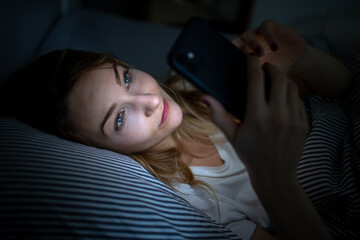 Young woman in bed holding a phone, tired and exhausted, blue light straining her eyes, messing up...