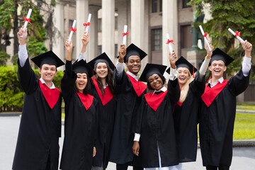 Cheerful multiracial students in graduation costumes raising diplomas up