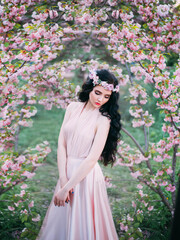 Naklejka premium Spring sexy woman standing in blooming sakura flowers garden. Medieval girl sad lady. pink silk dress. fairy tale image art photo. Dark long hair, wreath crown. cute face creative makeup. Green tree