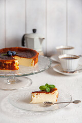 Slice of Homemade Basque burnt cheesecake on a plate with blueberries and mint leaves on light background, cups of coffee and geyser coffee maker
