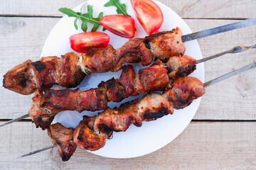 Juicy meat on skewers with tomatoes and herbs on a white plate on a wooden background. Kebab of meat is fried on the grill.