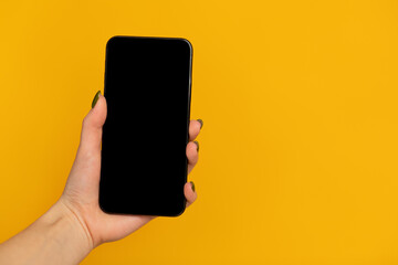 Fototapeta na wymiar Female hand holding smartphone with black blank screen for mockup on yellow studio background with free space