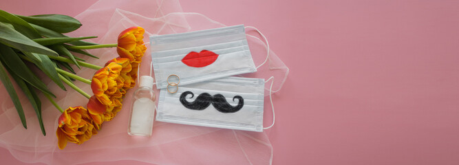 Two disposable protective masks with painted mustache and lips, two wedding rings and sanitizer next to bouquet on white veil on pink background. The wedding ceremony during COVID-19. Copy space