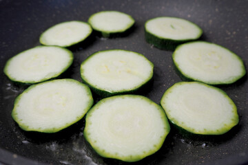 the process of frying green sliced zucchini slices in vegetable oil in a gray frying pan side view . vegetarianism . nine slices of zucchini