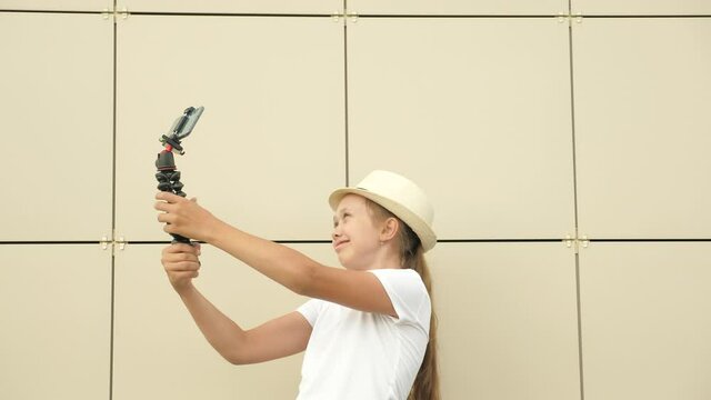 Modern girl in hat makes selfie outdoors. Fashionable teenager is photographed on smartphone using small tripod. Free girl blogger is conducting an online video conference from city street.