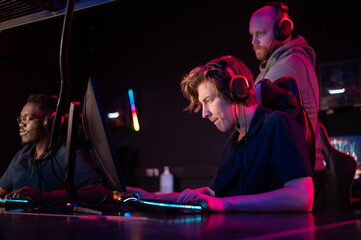 During the Dota 2 tournament, two guys from the team play at the computers, the coach watches from behind