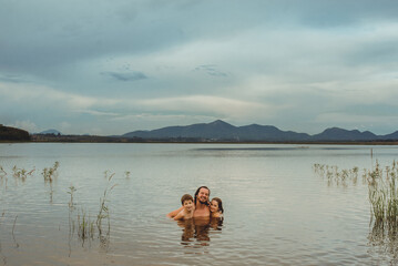 Obraz na płótnie Canvas Father hugging two kids (a boy and a girl) smiling into a calm lake, at Iraí Reservoir, in the city of Pinhais, State of Paraná, Brazil. In the background, the mountains that form the 