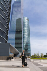 Mujer rubia ejecutiva caminando con maleta y con rascacielos de fondo en el centro de negocios