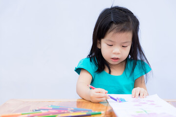 Girl in green shirt was sitting and coloring it with wood paint on drawing paper. Child 3-4 years old have fun making crafts. Kid do art on wooden table, train their imagination. White wall background
