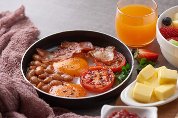 English breakfast in cooking pan with fried eggs, bacon, beans, grilled tomatoes. toast, butter, jam on a wooden board, orange juice, fresh fruits apples, blueberries, strawberries on stone background