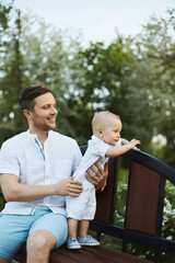 Fototapeta premium A handsome young man and adorable toddler boy, young dad and his little son, posing on the bench at a city park on a warm summer day