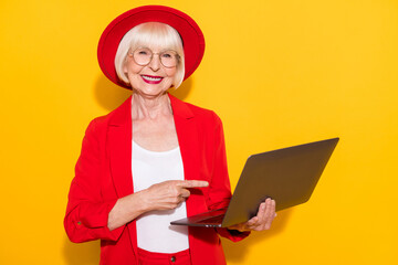 Photo portrait of old lady pointing on laptop wearing glasses red formal wear headwear isolated vivid yellow color background