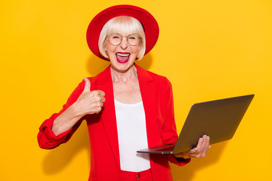 Photo Portrait Of Old Lady Wearing Red Suit Keeping Laptop Showing Thumb-up Sign Isolated Vibrant Yellow Color Background