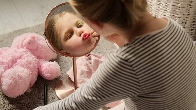 Little child girl paints her lips with lipstick, plays makes learn makeup role playing in front of the mirror in the children's room. Imitates adults making experiments with cosmetics.