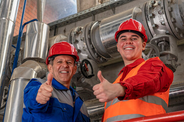Smiling Oil and Gas Engineer and Young Worker in Red Coveralls and Hardhat Giving Thumbs Up Beside Industrial Piping