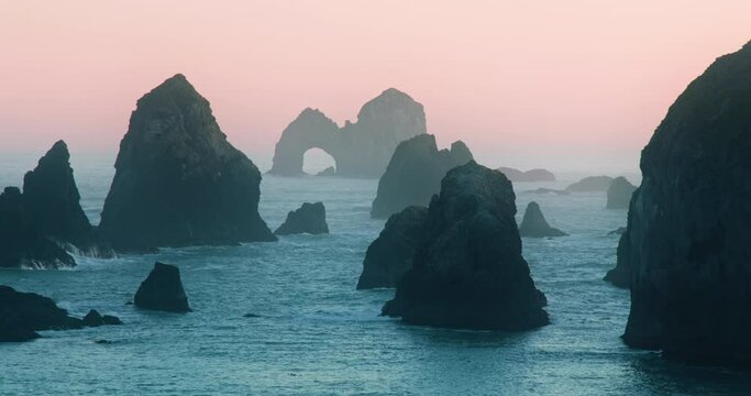 Cinematic sunset over rocky coast. Establishing shot of ocean nature landscape 4K footage. Wild nature park in pink sunset light. Blue ocean water surrounding scenic sea cliffs. Natural arch in rock