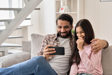 Happy indian father with teenage child daughter having fun using smart phone at home. Smiling dad and teen kid girl watching funny video, laughing at mobile apps sitting on sofa in living room.
