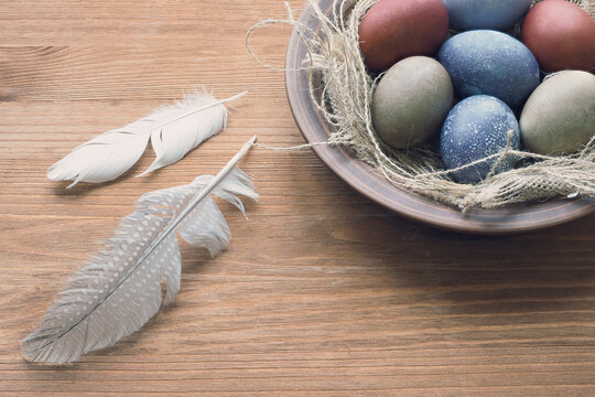 Ostara multicolored eggs in clay plate and two bird feathers on wooden background, close up, top view.