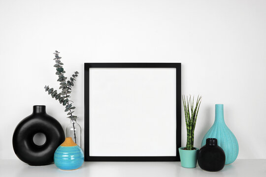 Mock up square black frame with houseplant, branches, black and blue decor. White shelf against a white wall. Copy space.