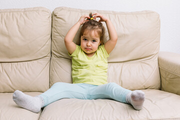 A girl with blond hair, sitting on a cream-colored sofa in a yellow shirt and blue pants, touching her head
