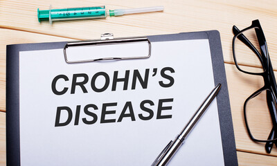 The words CROHN IS DISEASE is written on a white piece of paper next to black-rimmed glasses, a pen and a syringe. Medical concept