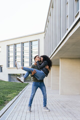 Happy afro couple having fun in the street. Latino boy carries his black-skinned girl in his arms between university buildings. Fun concept. Vertical photography.