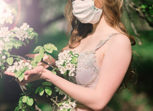 Beautiful young bride in a wedding dress and a white medical mask on her face near a blooming apple.