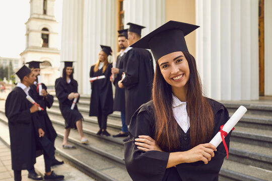 Portrait of a happy smiling graduate girl dressed in a black mantle with a diploma in her hands on the background of her classmates. Student poses on the steps of the university.