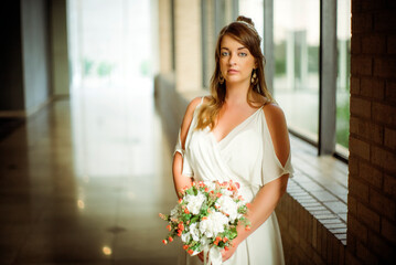 Beautiful young bride with flower bouquet standing to right of frame in large hall with windows  shallow depth of field