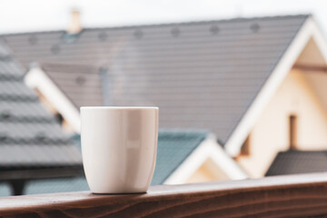 The steam rises from a cup of hot coffee or tea on the balcony with mountains and roofs of houses on the background. 