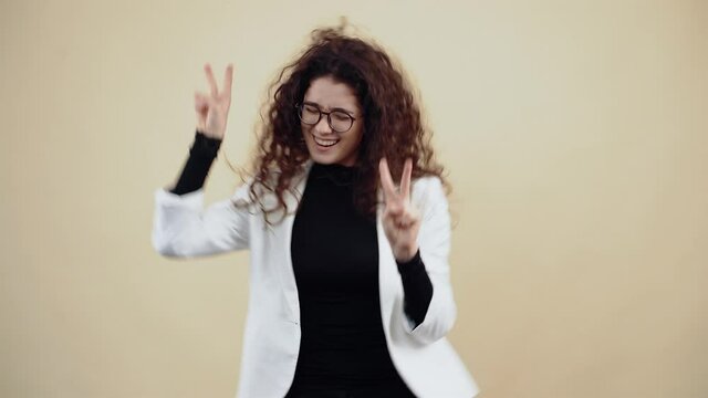 The distracted young woman with curly hair dances with her fingers in the shape of a peace sign, shaking her hands. Young hipster in gray jacket and white shirt, with glasses posing isolated on beige