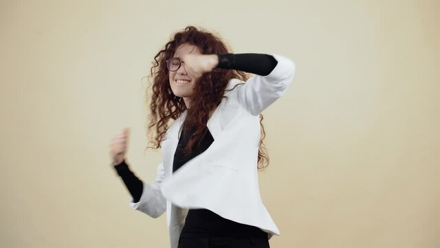 The young woman with curly hair dances with her hands, holds her fingers in her fists, shakes her hands to the left and to the right, fooling around. Young hipster in gray jacket and white shirt,
