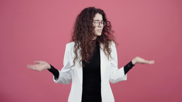 The young woman is confused with her curly hair, gestures with her hands and does not understand why, she had moved from side to side. Young hipster in white jacket and black shirt, with glasses
