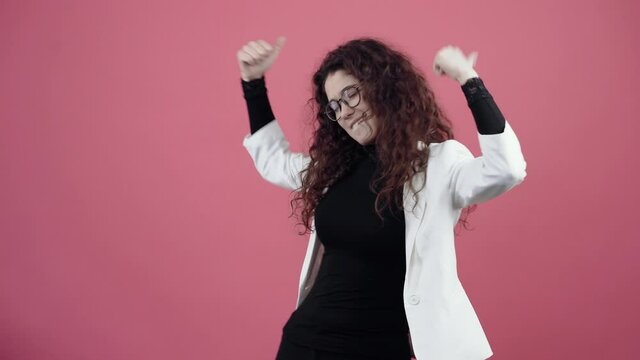 The happy young woman with curly hair dances with her hands, two open fingers fluttering. Young hipster in white jacket and black shirt, with glasses posing isolated on pink background in the studio.