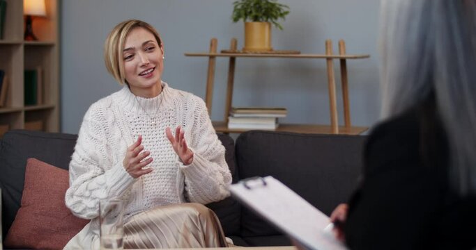 Crop view of cheerful woman with short blond hair talking with therapist in office . Female pretty person communicating with psychologist while sitting on sofa at therapy session.