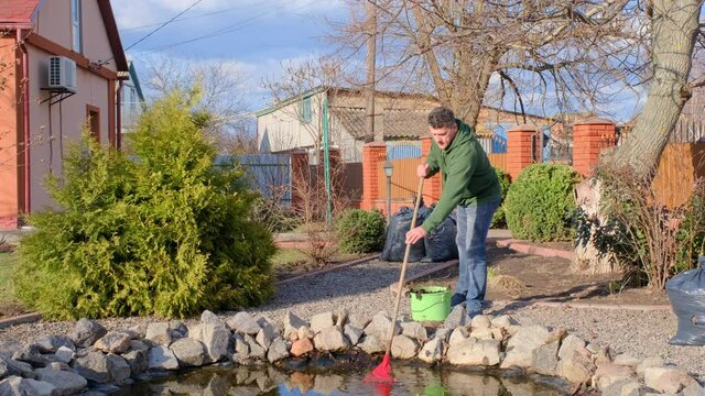 Middle-aged caucasian man cleans a garden pond with leaf rake from water plants and falling leaves near his house. Spring seasonal pond care after winter. 