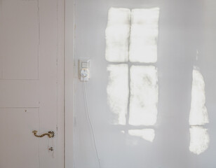 Die Sonne projiziert ein Fenster auf die weiße Wand eines Zimmers mit alter lackierter Tür und Lichtschalter mit Steckdose
