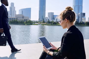 Young blond elegant businesswoman in formalwear using tablet while sitting by riverside in urban environment with man passing by